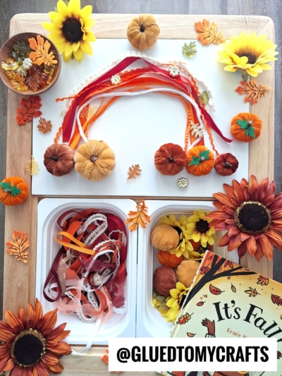 A fall-themed craft setup with orange and yellow flowers, small pumpkins, colorful ribbons, faux leaves, and a “It’s Fall” book arranged on a table—perfect inspiration for FALL THEMED SENSORY BIN IDEAS. Supplies are organized below; @GLUEDTOMYCRAFTS labeled.
