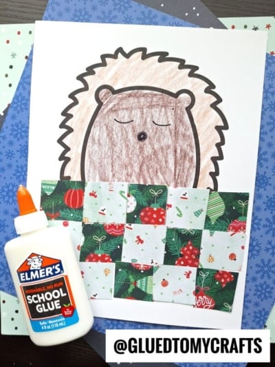 A sleeping hedgehog craft features a cozy hedgehog under a patchwork quilt made of holiday-patterned paper squares. Elmer’s school glue sits beside the artwork, surrounded by colorful papers in the background.