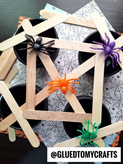 Four black cups on a spiderweb-patterned paper, topped with craft stick webs and colorful toy spiders in black, orange, purple, and green. Perfect for fall themed sensory bin ideas! A label at the bottom reads @GLUEDTOMYCRAFTS.