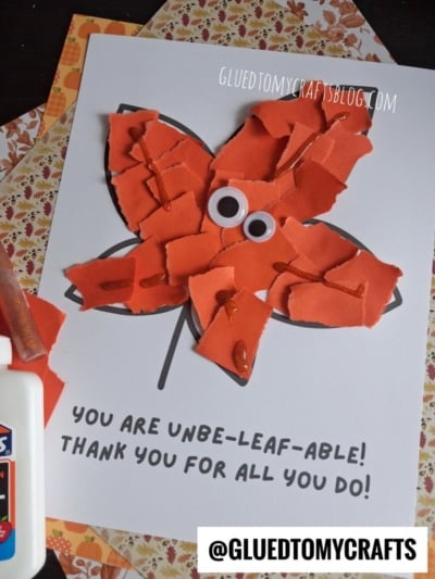A card features a torn paper leaf in bright orange with googly eyes, outlined in black, on a white background. Text reads: “You are unbe-leaf-able! Thank you for all you do!” Glue and patterned paper are nearby.