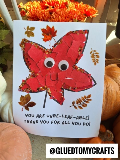 A handmade card features a large red torn paper leaf with googly eyes and a smile, decorated with glitter and real leaves. Text reads, “You are unbe-leaf-able! Thank you for all you do!” Fall pumpkins and flowers surround the card.