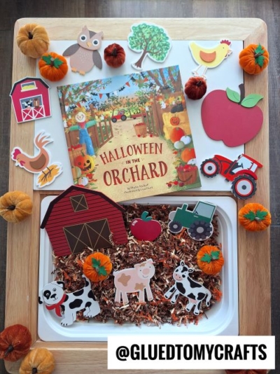 A Halloween-themed sensory tray, perfect for FALL THEMED SENSORY BIN IDEAS, features toy cows, a barn, pumpkins, a tractor, and the book Halloween in the Orchard—all surrounded by apple and owl cutouts. Username @GLUEDTOMYCRAFTS is visible.