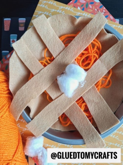 A craft project resembling a pumpkin pie, perfect for FALL THEMED SENSORY BIN IDEAS, made with orange yarn, tan felt strips as a crust, and cotton balls as whipped cream, sits on a fall-themed background. The @GLUEDTOMYCRAFTS handle is visible.