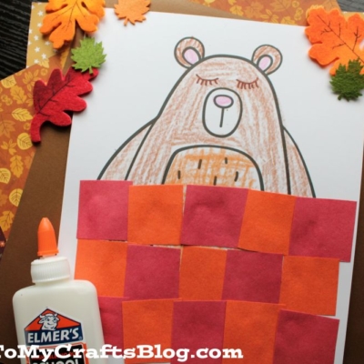 A hibernating bear craft featuring a brown bear sleeping under a red and orange paper blanket. Elmer’s School Glue, felt leaves, and paper leaves decorate the scene.
