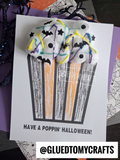 Halloween popcorn card featuring a black-and-orange striped container drawn on the front, topped with fluffy cotton balls for popcorn, googly eyes, painted lines, and festive bat confetti. Text reads, Have a Poppin Halloween! and @GLUEDTOMYCRAFTS.