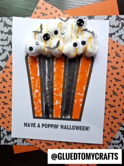 Handmade halloween popcorn card featuring a popcorn tub made of orange and black stripes, topped with fluffy cotton balls, googly eyes, and plastic spiders. Text reads, Have a poppin Halloween! Card rests on bat-patterned paper.