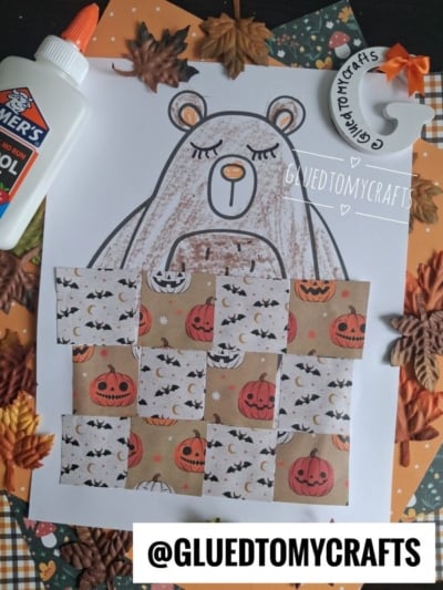 A hibernating bear craft outline colored in, decorated with squares of Halloween-themed paper, is surrounded by fall leaves, glue, and orange craft paper. Text @GLUEDTOMYCRAFTS appears at the bottom.