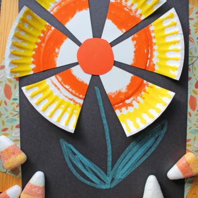 A paper plate candy corn flower is crafted by cutting and painting a plate with orange and yellow petals, then arranging it on black paper with a drawn green stem and leaves, surrounded by real candy corn pieces.