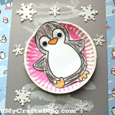 A handmade snow tubing penguin craft features a paper plate, colored paper, and drawn details as it slides on gray paper with chalk streaks and paper snowflakes.