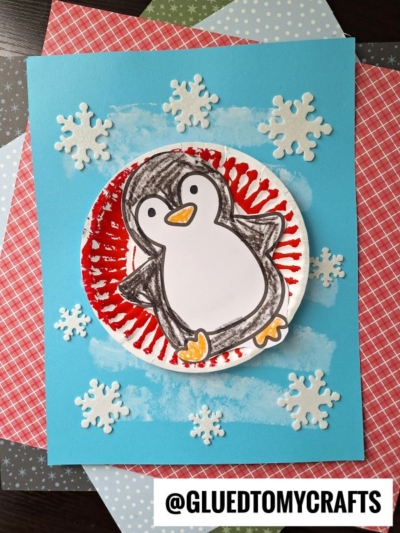 A handmade snow tubing penguin craft on blue paper, featuring a colorful penguin on a red and white circle, surrounded by white snowflakes and layered patterned papers. Text at the bottom reads @GLUEDTOMYCRAFTS.