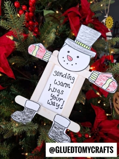 A popsicle stick snowman picture frame ornament made of paper and wood sits on a Christmas tree. The snowman holds a sign that says, sending warm hugs your way! Red poinsettias decorate the tree. @GLUEDTOMYCRAFTS appears at the bottom.