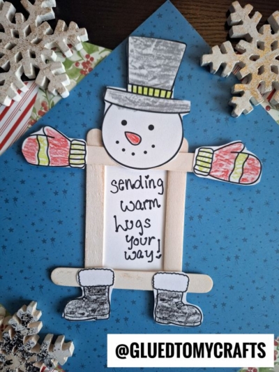 A handmade popsicle stick snowman picture frame craft, featuring red mittens and gray boots, sits on a blue background with snowflakes. A note on its body reads, sending warm hugs your way!.