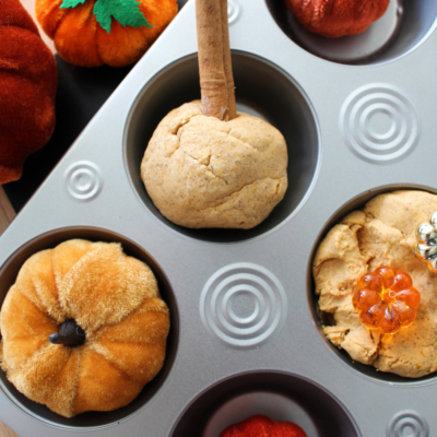 A muffin tin holds a Pumpkin Spice Cloud Dough ball with a cinnamon stick stem, surrounded by plush and decorative pumpkin ornaments. The image highlights the cozy fun of making Pumpkin Spice Cloud Dough crafts.