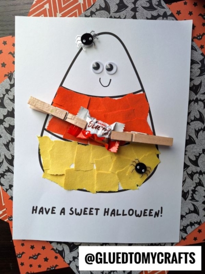 A torn paper candy corn craft made with colorful ripped paper pieces, googly eyes, a big smile, plastic spiders, and a mini candy held by a clothespin. Text says, HAVE A SWEET HALLOWEEN! and @GLUEDTOMYCRAFTS at the bottom.