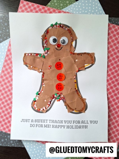 A handmade torn paper gingerbread man craft with googly eyes, red buttons, and glitter on brown paper, glued to a white sheet with a Happy Holidays message. Elmers glue and Christmas-patterned paper are nearby.