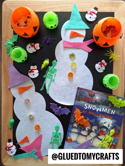 A Halloween-themed craft tray featuring felt snowman shapes, mini pumpkins, spiders, bats, toy skeletons, and the book “Snowmen at Halloween”—perfect for FALL THEMED SENSORY BIN IDEAS that spark creativity and festive fun!.