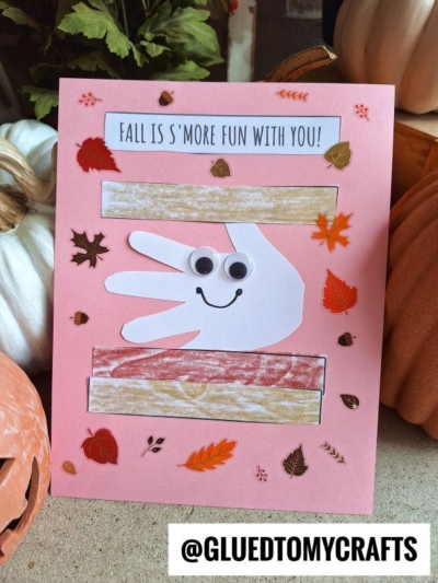 A pink card features a handprint s’mores keepsake made with paper strips like graham crackers and chocolate, googly eyes, a smile, and fall leaf stickers. Text at the top says, “Fall is s’more fun with you!”.