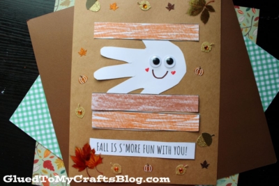 A fall-themed craft features a handprint smores keepsake decorated with googly eyes and a smile, surrounded by leaf and pumpkin stickers. Text reads, “Fall is s’more fun with you!”