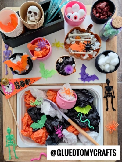 A Halloween-themed sensory bin with black and orange tissue, plastic spiders, bats, skeletons, fake eyeballs, cotton balls, and toy bones—perfect for those seeking fall themed sensory bin ideas! A sign reads @GLUEDTOMYCRAFTS.