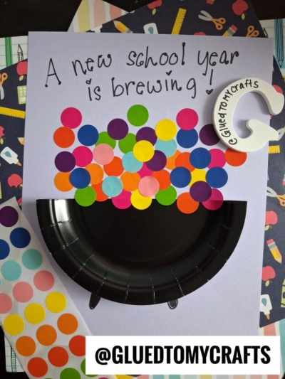 A craft project with a purple paper reading A new school year is brewing, showcasing a paper plate bubbling cauldron and colorful dot stickers as bubbles. Sticker sheets and a glue ring sit nearby. @GLUEDTOMYCRAFTS appears at the bottom.