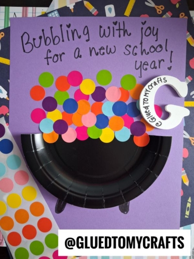A purple paper displays a paper plate craft resembling a bubbling cauldron, with colorful dot stickers as gum balls. Bubbling with joy for a new school year! is written at the top. @GLUEDTOMYCRAFTS is at the bottom.