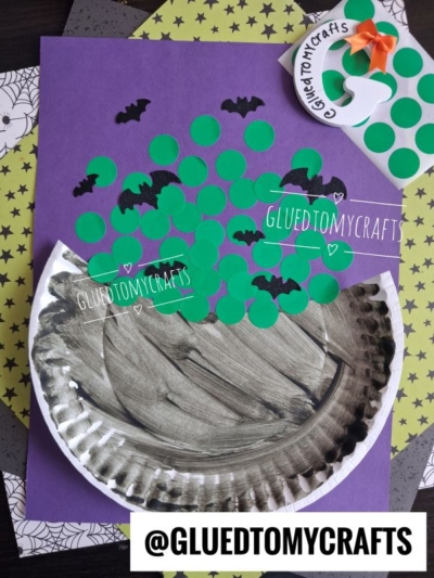 A Halloween craft shows a paper plate bubbling cauldron, painted black and decorated with green paper dots to look like potion bubbles, along with black bat cutouts. The spooky scene is set on a purple background with spiderweb-patterned papers.