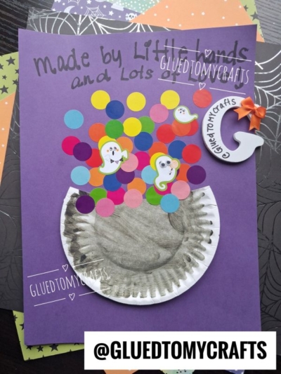 A purple craft paper features a painted paper plate bubbling cauldron, colorful paper dots, ghost stickers, and a small paper moon with an orange bow. The text @GLUEDTOMYCRAFTS appears at the bottom.