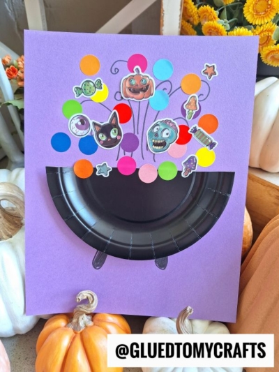 A Halloween craft featuring a paper plate bubbling cauldron made from a black paper plate on purple paper, decorated with colorful dots and Halloween-themed stickers like a pumpkin, black cat, and skull. Text reads @GLUEDTOMYCRAFTS.
