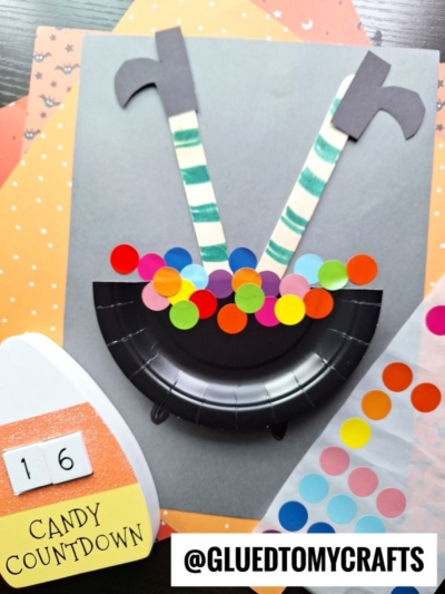 A Halloween craft features a paper plate bubbling cauldron with colorful paper bubbles, striped witch legs, and pointy shoes. Behind is orange polka-dot paper, a candy corn cutout labeled 16 Candy Countdown, and the @GLUEDTOMYCRAFTS handle.