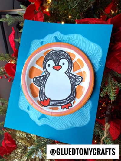 A handmade craft featuring a snow tubing penguin drawn on a paper plate, attached to a blue paper background with an ice-like effect, displayed on a Christmas tree. The image has the text @GLUETOMYCRAFTS at the bottom.