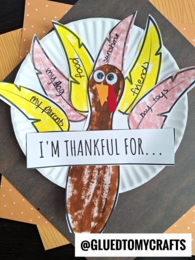 A paper plate thankful turkey craft featuring vibrant feather cutouts labeled with gratitude notes like “my dog,” “my parents,” and “sunshine.” A cheerful sign reads IM THANKFUL FOR... above this creative Thanksgiving project.