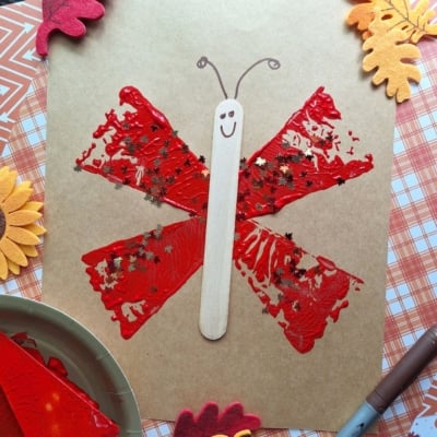 A butterfly craft made with red paint stamped in wing shapes using a pie server on brown paper, a popsicle stick as the body with a drawn smiley face, and star-shaped glitter. Fall-themed felt leaves and craft supplies surround the artwork.
