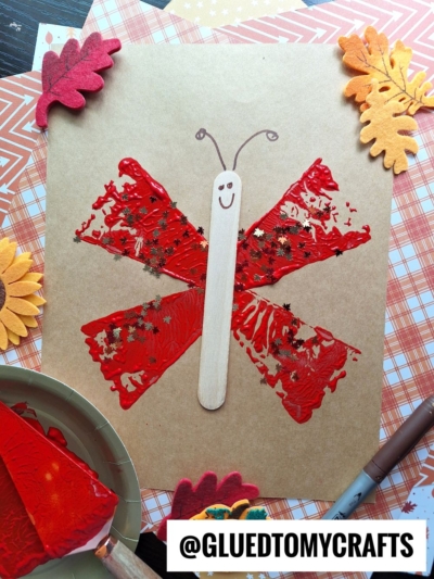 A butterfly craft made with red paint stamped in wing shapes using a pie server on brown paper, a popsicle stick as the body with a drawn smiley face, and star-shaped glitter. Fall-themed felt leaves and craft supplies surround the artwork.