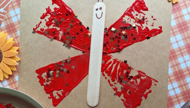 A butterfly craft made with red paint stamped in wing shapes using a pie server on brown paper, a popsicle stick as the body with a drawn smiley face, and star-shaped glitter. Fall-themed felt leaves and craft supplies surround the artwork.