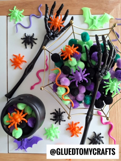 Halloween-themed craft supplies, perfect for FALL THEMED SENSORY BIN IDEAS, include black skeletal hands, colorful pom-poms, plastic spiders, bats, and a cauldron. A spiderweb tray holds a pile of green, purple, orange, and black pom-poms.
