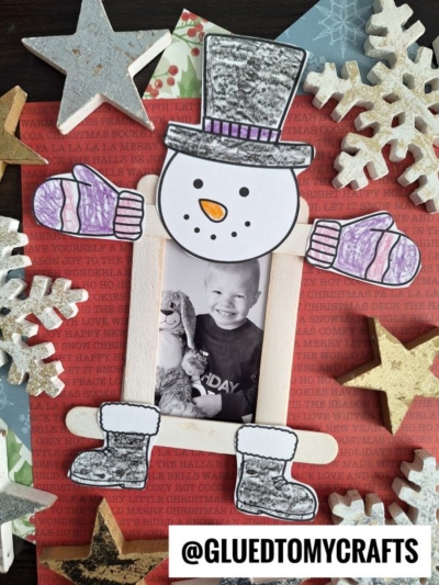 A handmade popsicle stick snowman picture frame decorated with a child’s photo, mittens, hat, and boots, surrounded by stars and snowflakes on a festive background. The Instagram handle @GLUEDTOMYCRAFTS is visible at the bottom.