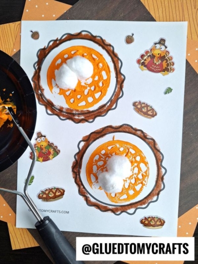 A Thanksgiving craft page features two Potato Masher Stamped Pumpkin Pie drawings decorated with orange paint and cotton balls, surrounded by turkey and pumpkin pie stickers. A paint roller and orange paper are also visible. @GLUEDTOMYCRAFTS is labeled at the bottom.