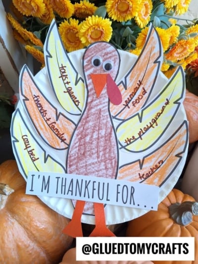 A vibrant paper plate thankful turkey features handwritten notes of gratitude—like family, playground, food, teachers, and cousins—on its feathers, surrounded by pumpkins and yellow flowers.