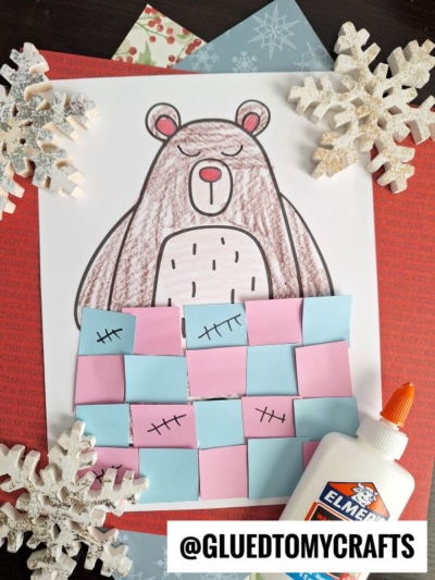 A drawing of a brown bear tucked under a paper quilt made of pink and blue squares, surrounded by snowflake decorations, a bottle of glue, and patterned paper. This cozy hibernating bear craft features the handle @GLUEDTOMYCRAFTS at the bottom.