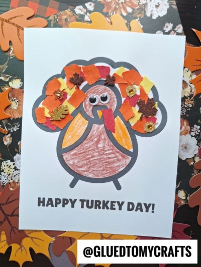 A child’s torn paper turkey craft with colorful paper leaf feathers, googly eyes, and HAPPY TURKEY DAY! text, placed on a table decorated with autumn leaf cutouts.