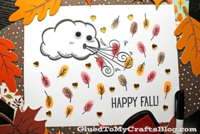 Autumn-themed craft page with a cartoon wind cloud blowing thumbprint leaves, googly eyes, and gold heart stickers. Surrounded by paper leaves and ink pads, with text reading HAPPY FALL!