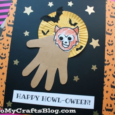 A Halloween craft featuring a handprint werewolf made from a brown paper hand as the wolf’s body, a paper wolf face, and a yellow moon with bats on a black background, surrounded by gold stars. Text reads HAPPY HOWL-OWEEN!