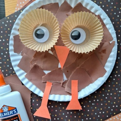 Paper plate owl craft made with brown paper pieces, cupcake liners for eyes, googly eyes, and orange paper for beak and feet—perfect for FALL CRAFTS FOR KIDS. Elmers glue and patterned paper are in the background. @GLUEDTOMYCRAFTS is at the bottom.