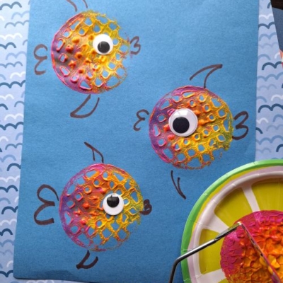 A blue sheet of paper displays three colorful fish made from painted bubble wrap circles, each with a googly eye and black marker details—perfect for pairing with a potato masher stamped craft. Art supplies and patterned paper fill the background.