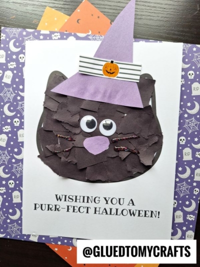 A Halloween craft showcases a charming torn paper black cat with googly eyes and a whimsical purple witch hat, all set on white paper that reads “Wishing you a purr-fect Halloween!” The creation is finished off with the @GLUETOMYCRAFTS tag at the bottom.