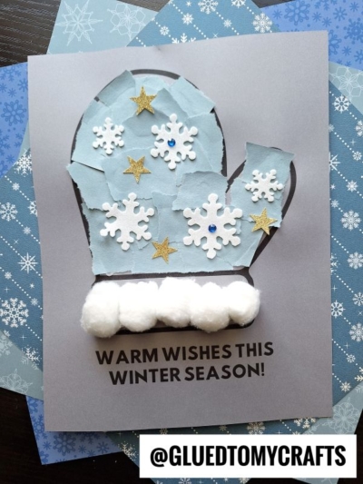 A torn paper mitten craft decorated with white snowflakes, gold stars, blue gems, and white pom-poms, on a gray card that says, Warm wishes this winter season! The background has blue snowflake patterns.