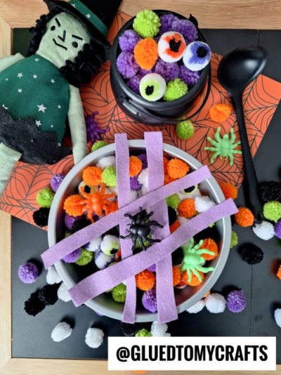 A Halloween craft setup featuring a witch doll, bowls filled with colorful pom-poms and toy spiders—one covered with purple felt strips like a web. Perfect for FALL THEMED SENSORY BIN IDEAS, with Halloween-themed paper and scattered pom-poms completing the scene.