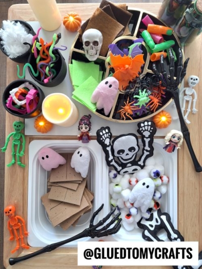 A Halloween-themed craft and snack setup features felt ghosts, skeleton cutouts, toy figurines, fake candles, colored felt, marshmallows, candy, and crackers—perfect inspiration for FALL THEMED SENSORY BIN IDEAS. Text: @GLUEDTOMYCRAFTS.