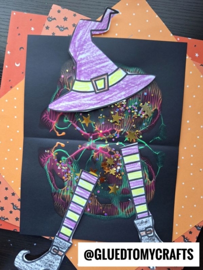 A Halloween craft features a purple witch hat, striped witch boots, and glitter on a black paper jack-o-lantern set on orange star-patterned paper, with a playful paint splat melted witch effect adding spooky charm. @GLUEDTOMYCRAFTS is displayed at the bottom.