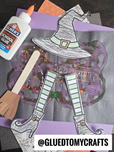 Craft supplies and witch-themed cutouts, including a hat, shoes, and striped legs that appear like a paint splat melted witch, are arranged on colored paper alongside Elmers glue, a wooden stick, and glitter. GLUEDTOMYCRAFTS watermark is visible.